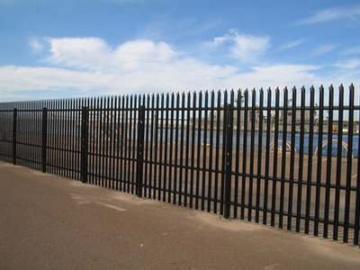 A long row of black powder coated palisade fencing around the sea and there is a ship in the sea.
