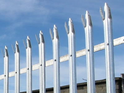 A part of high galvanized palisade fence from below, the fence head is triple pointed and installed next to the brick wall.